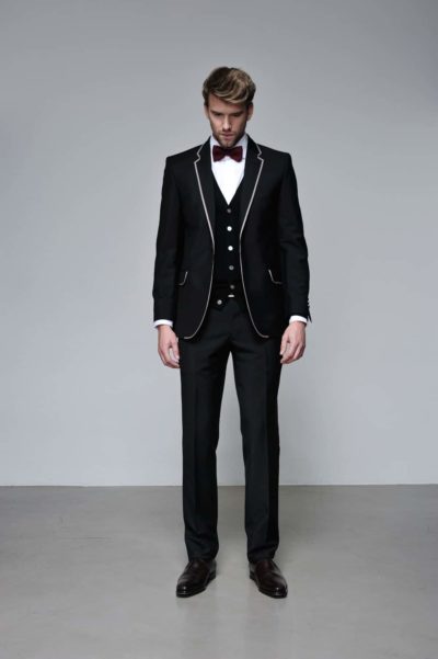 Un mannequin en smoking noir avec bordure blanche et gilet assorti se tient debout sur un fond gris uni. Il porte une chemise blanche, un nœud papillon violet et des chaussures noires formelles. Ses cheveux sont soigneusement coiffés et il regarde vers le bas avec une expression sérieuse, capturée par le photographe Thomas Duclert à Paris.