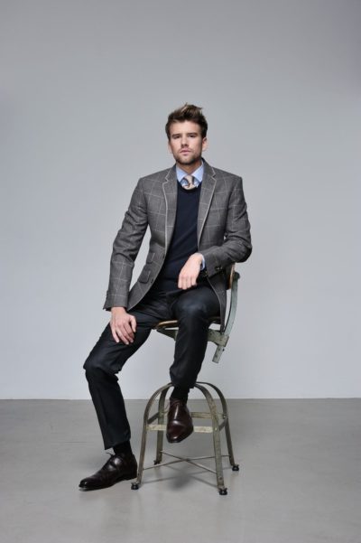 Un mannequin avec une barbe soignée et des cheveux coiffés est assis sur un tabouret. Il porte un blazer à carreaux gris sur un pull bleu marine et une chemise bleu clair, associés à un pantalon foncé et des chaussures marron. En mode, il semble confiant, posant comme s'il était prêt pour une photo à Paris sur le fond gris uni.