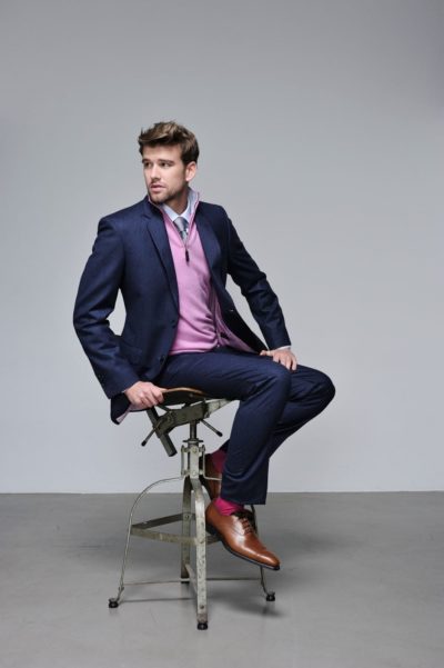 Un mannequin aux cheveux courts et à la barbe taillée est assis sur un tabouret en métal de style industriel. Vêtu d'un blazer bleu marine, d'un pantalon bleu et d'un pull rose zippé superposé sur une chemise bleu clair, il incarne la mode sophistiquée. Des chaussures marron complètent son look sur fond gris uni capturé par le photographe Thomas Duclert.