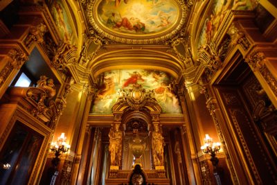 Une salle luxueuse et opulente avec des détails architecturaux dorés complexes, de grandes colonnes ornées et des peintures classiques au plafond rappelant l'élégance parisienne. Illuminée par la lumière chaleureuse des lustres et des appliques murales, la riche décoration n'est rien de moins que le plus raffiné des styles imaginés par Thomas Duclert.