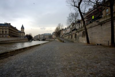 Une allée pavée longe une rivière à Paris, bordée d'arbres nus et de bâtiments historiques. Le ciel couvert donne une teinte grisâtre à la scène, tandis qu'un pont est visible en arrière-plan enjambant la rivière, capturant l'essence de la ville telle que le photographe Thomas Duclert pourrait la voir.