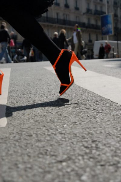 Une mannequin en bas noirs et chaussures à talons hauts orange vif, marchant sur un passage piéton, capture l'essence de la mode urbaine. Photographiée par Thomas Duclert, la scène met en scène des piétons animés et de grands immeubles sous un ciel dégagé.