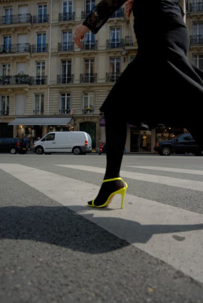 Une mannequin portant une longue jupe noire et des talons hauts jaunes traverse un passage piéton dans une rue de Paris, avec des bâtiments et des véhicules stationnés visibles en arrière-plan. L'accent est mis sur les jambes et les chaussures, capturant le mouvement, la vie urbaine et la mode parisienne.