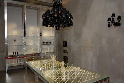 Une salle à manger moderne et minimaliste avec une longue table en verre éclairée entourée de chaises argentées. Le style de la pièce est accentué par un lustre noir et des appliques murales assorties. Des étagères avec de la verrerie bordent le mur et les murs en béton ajoutent une touche industrielle rappelant le chic parisien.