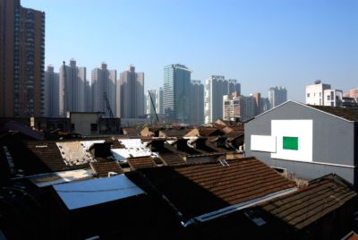Un paysage urbain qui contraste avec les vieux bâtiments bas aux toits de tuiles usés au premier plan et les immeubles d'appartements modernes de grande hauteur sous un ciel bleu clair à l'arrière-plan. Quelques grues sont visibles derrière les vieilles structures, indiquant une activité de construction, une scène qui rappelle Paris capturée par un photographe expérimenté.