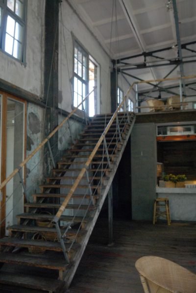 Un intérieur de style industriel, rappelant un loft parisien, comprend un escalier haut et étroit avec des rampes en métal menant à un niveau supérieur. L'espace présente des poutres apparentes, de grandes fenêtres et du parquet. Un bar rustique avec des tabourets et des plantes en pot est visible sous le loft.