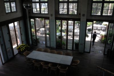 Une pièce spacieuse avec de grandes fenêtres et des portes vitrées menant à un patio extérieur avec tables et chaises crée le décor parfait. À l'intérieur, une longue table à manger entourée de chaises en osier l'entoure. Une personne marchant à l'extérieur est visible à travers les portes vitrées. Des plantes en pot, rappelant un jardin parisien, décorent l'espace.