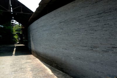 Une étroite allée pavée à Paris, bordée d'un haut mur de briques texturées sur la droite. La lumière du soleil crée des ombres tout au long du chemin, et le chemin s'étend dans une zone couverte avec de la verdure visible en arrière-plan, une scène idéale pour un photographe de mode.