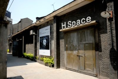 Vue extérieure d'un bâtiment aux murs de briques sombres et au-dessus d'une grande enseigne portant l'inscription « H Space » (Espace H) à double battant en bois. Une affiche présentant des œuvres monochromes est exposée à gauche de l'entrée, évoquant la mode parisienne. Des plantes vertes bordent le mur extérieur du bâtiment, parfaites pour l'objectif de tout photographe.