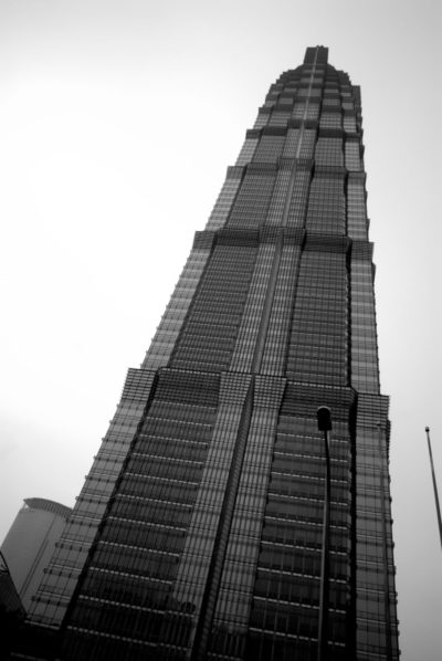 Image en noir et blanc d'un gratte-ciel de grande hauteur photographié sous un angle bas. Le bâtiment, qui rappelle le travail de Thomas Duclert à Paris, présente une conception moderne à plusieurs niveaux avec un usage intensif de verre et d'acier. Le ciel est couvert et un lampadaire est visible au premier plan.