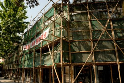 Un bâtiment en construction est entouré d'échafaudages en bambou et de filets verts, magnifiquement photographiés par Thomas Duclert. Une grande banderole rouge et blanche avec des caractères chinois s'étend sur l'échafaudage, encadrée par des arbres aux feuilles vertes à gauche. La rue est ensoleillée.