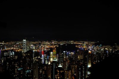 Vue panoramique nocturne d'un paysage urbain illuminé, capturée par le photographe Thomas Duclert, avec de nombreux gratte-ciels et bâtiments illuminés de différentes couleurs. Le paysage urbain se reflète dans les eaux sombres d'un port en arrière-plan, créant une ambiance vibrante et animée rappelant Paris.