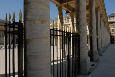 Une scène architecturale classique avec une rangée de grandes colonnes en pierre et des portes en fer noir avec des pointes à pointes dorées sous une colonnade ombragée. Photographié par Thomas Duclert, le décor de bâtiments historiques sur fond de ciel bleu suggère un cadre parisien pittoresque.