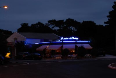 Un petit restaurant nommé "Le P'tit Chalet" est illuminé par des néons bleus le soir. Des tables et des chaises d'extérieur sont installées sous un auvent, et quelques voitures sont garées à proximité. Le photographe Thomas Duclert a capturé cette scène tranquille, rappelant un coin tranquille de Paris, entouré d'arbres.