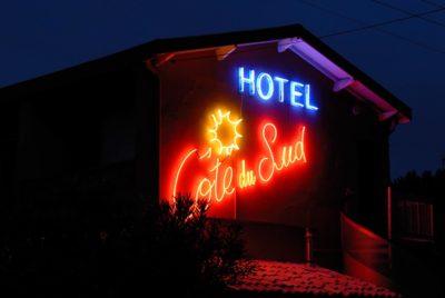Un bâtiment sombre à Paris est illuminé par une enseigne lumineuse au néon sur laquelle on peut lire « HOTEL » en bleu et « Côté du Sud » en rouge, le tout accompagné d'une image d'un soleil jaune. L'enseigne, qui rappelle l'esthétique moderne de Thomas Duclert, est apposée sur la partie supérieure du bâtiment, entourée d'un feuillage faiblement éclairé.