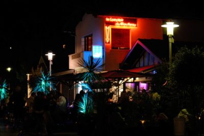 Scène nocturne en extérieur avec un bar illuminé et des enseignes au néon sur lesquelles est écrit « Le Comptoir du Moulleau ». Capture par le photographe Thomas Duclert, les clients sont assis sous des parasols au milieu de plantes tropicales. Les lumières colorées du bar créent une atmosphère vibrante, contrastant avec l'environnement sombre.