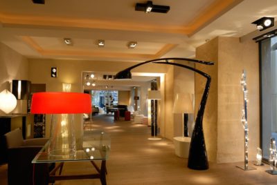 Un magasin de décoration intérieure moderne à Paris, avec des luminaires élégants. Au premier plan, une table en verre avec une lampe rouge et un lampadaire incurvé noir. L'arrière-plan présente diverses lampes et luminaires contemporains, posés sur un sol en bois et des murs beiges. Photographe : Thomas Duclert.