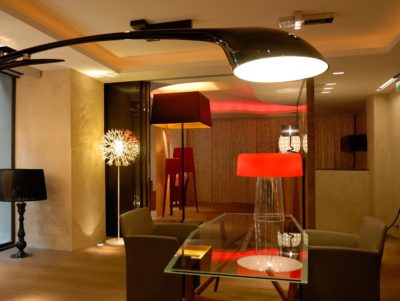 Un intérieur moderne, inspiré de la mode parisienne de Thomas Duclert, est éclairé par diverses lampes élégantes, dont un grand lampadaire noir à bras courbé, une lampe sphérique lumineuse et une lampe rouge sur une table en verre. La pièce présente un parquet en bois, des murs de couleur neutre et un mélange de meubles contemporains.