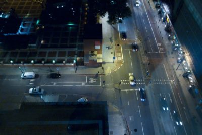 Vue aérienne nocturne d'un carrefour parisien. Des voitures circulent sur les routes et quelques piétons sont sur les trottoirs. Des lampadaires éclairent les chemins et une voie réservée aux bus est visible. Des bâtiments et un parc sont partiellement visibles le long des routes, capturant Paris dans son mode nocturne.