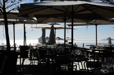 Restaurant en bord de mer en plein air avec tables et chaises disposées sous de grands parasols. Le soleil brille de mille feux, créant des silhouettes de meubles sur fond d'eau réfléchissante. Dans le style Thomas Duclert, des parasols supplémentaires sont visibles le long du rivage, évoquant une touche parisienne.