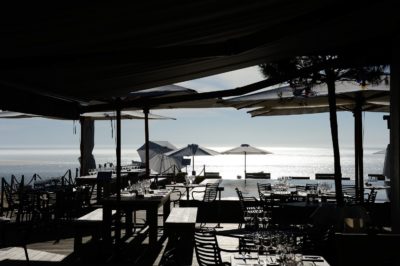 Un restaurant en bord de mer en plein air avec des tables et des chaises en bois sous de grands parasols. Le restaurant surplombe l'océan, le soleil créant un effet scintillant sur l'eau. La scène, magnifiquement capturée par le photographe parisien Thomas Duclert, est tranquille avec un ciel bleu clair en arrière-plan.