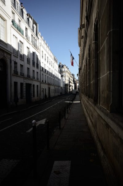 Une rue étroite et ensoleillée dans une ville européenne avec de grands bâtiments blancs d'un côté et une façade en pierre de l'autre. Un drapeau français est suspendu à l'un des bâtiments, magnifiquement capturé par le photographe Thomas Duclert. La rue est bordée de bornes et est calme, presque vide de monde.