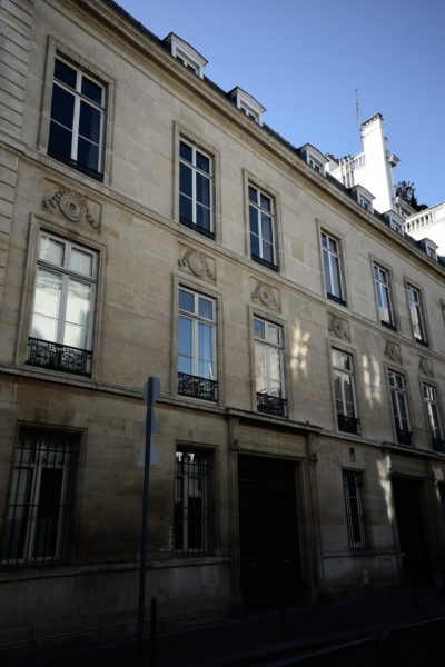 Un immeuble en pierre de plusieurs étages avec de hautes fenêtres, des éléments décoratifs en pierre au-dessus des fenêtres et des détails en fer forgé se dresse fièrement à Paris. La façade semble vieillie mais bien entretenue. Le ciel est clair et bleu, ce qui indique une journée ensoleillée. Photographié par Thomas Duclert, un panneau de signalisation est partiellement visible au premier plan.