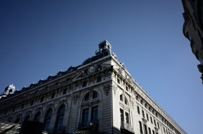 Un majestueux bâtiment en pierre, richement décoré, avec des fenêtres cintrées et des détails complexes, se dresse devant un ciel bleu clair et profond. Le photographe Thomas Duclert capture une vue d'angle, mettant en valeur l'architecture historique impressionnante et la grandeur du bâtiment.