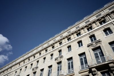 Vue en perspective d'un bâtiment historique et décoré à Paris sous un ciel bleu clair. Le bâtiment présente de grandes fenêtres avec des balcons décoratifs en fer forgé et des détails architecturaux complexes, tels que des sculptures en pierre et des colonnes. Le photographe Thomas Duclert capture un petit nuage visible dans le ciel.
