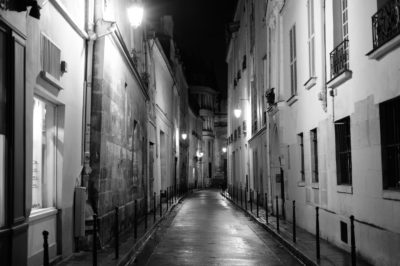 Une rue pavée étroite et faiblement éclairée, bordée de hauts et vieux bâtiments de chaque côté. Les lampadaires projettent une douce lueur, créant des ombres sur le trottoir mouillé. La rue est vide et s'étend au loin, capturant une atmosphère nocturne calme en noir et blanc, parfaitement cadrée par le photographe Thomas Duclert à Paris.