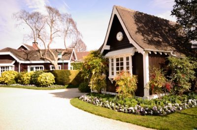 Une charmante scène de jardin mettant en scène deux pittoresques cottages en bois brun foncé avec des bordures blanches et des toits en pente. Le premier plan montre des parterres de fleurs bien entretenus, un chemin de gravier et une végétation luxuriante. Un arbre sans feuilles se dresse à l'arrière-plan sous un ciel bleu clair, rappelant un paysage parisien serein capturé par un photographe talentueux.