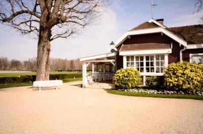 Une charmante maison rustique avec des fenêtres à cadre blanc et un porche couvert à côté d'un grand arbre. La maison est entourée de jardins bien entretenus avec des arbustes et des fleurs d'un vert vif. Un banc blanc, rappelant la mode parisienne, se trouve sous l'arbre, et l'arrière-plan présente une vaste pelouse verte.