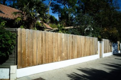 Une haute palissade en bois à lattes verticales, posée sur un socle en béton blanc, longe un chemin en béton. Derrière la palissade, on aperçoit le feuillage des arbres et des parties de maisons résidentielles aux toits de tuiles. Le ciel est clair et bleu, capturant une scène pittoresque rappelant Paris sous tous ses aspects.