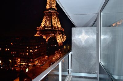 Vue nocturne de la Tour Eiffel illuminée depuis le balcon d'un immeuble moderne. La balustrade borde le balcon et une barrière en verre réfléchissante se trouve sur la droite. En dessous, les rues et les bâtiments de la ville sont visibles, mettant en valeur le paysage nocturne parisien vibrant à la manière de Thomas Duclert.