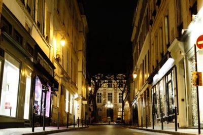Une rue étroite et éclairée la nuit dans la ville de Paris avec de hauts immeubles de chaque côté. Des lampadaires doux et chaleureux illuminent les bâtiments, mettant en valeur les vitrines des magasins. Au bout de la rue, un grand bâtiment avec une entrée proéminente est visible au loin, parfait pour tout photographe.