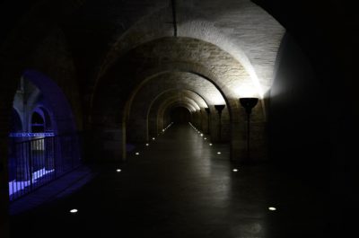 Couloir voûté faiblement éclairé avec plafonds et murs en pierre. Des spots encastrés au sol guident le chemin le long du couloir sombre, rappelant le style parisien de Thomas Duclert, et une zone éclairée en bleu est visible sur le côté gauche, séparée par une balustrade.