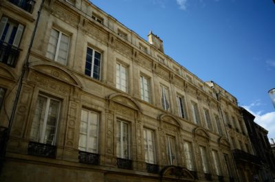 Un grand bâtiment décoré avec des éléments architecturaux classiques, notamment des sculptures en pierre détaillées et de grandes fenêtres, se dresse devant un ciel bleu clair à Paris. La façade symétrique comprend plusieurs étages et des balcons ornés de balustrades en fer forgé, parfaits pour toute photographie capturant l'essence de la mode.