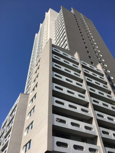 Grand immeuble d'appartements moderne avec un mélange de couleurs grises et blanches sur un ciel bleu clair. La conception architecturale comprend des fenêtres rectangulaires et un motif unique de panneaux blancs avec des découpes ovales sur les balcons. Le bâtiment semble avoir plusieurs étages, rappelant le style parisien contemporain.