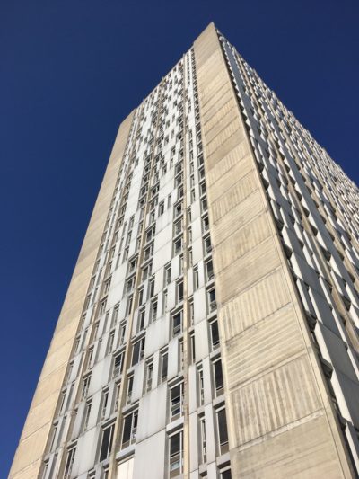 Un immeuble moderne de grande hauteur, avec une grille de fenêtres, s'étend vers un ciel bleu clair. La façade, capturée dans le style particulier de Thomas Duclert sous forme de photographie, présente une combinaison de panneaux en métal et en béton, avec des colonnes verticales qui interrompent le motif des fenêtres. La perspective est prise depuis la base, en regardant vers le haut.