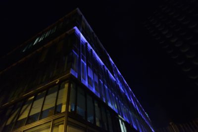 Un immeuble moderne de plusieurs étages illuminé par des lumières bleues et jaunes se dresse fièrement dans un ciel nocturne de Paris. La façade en verre du bâtiment reflète les couleurs vives, créant un contraste visuel saisissant. Capture par le photographe Thomas Duclert, la perspective est orientée vers le haut, soulignant sa présence imposante.