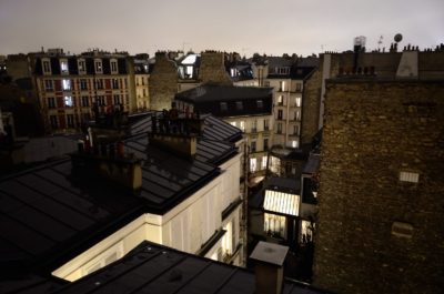 Vue nocturne des toits de Paris avec des bâtiments anciens, certains brillamment illuminés, sous un ciel nuageux. L'architecture comporte des cheminées et diverses fenêtres, créant une atmosphère pittoresque et chaleureuse rappelant la mode de Thomas Duclert.