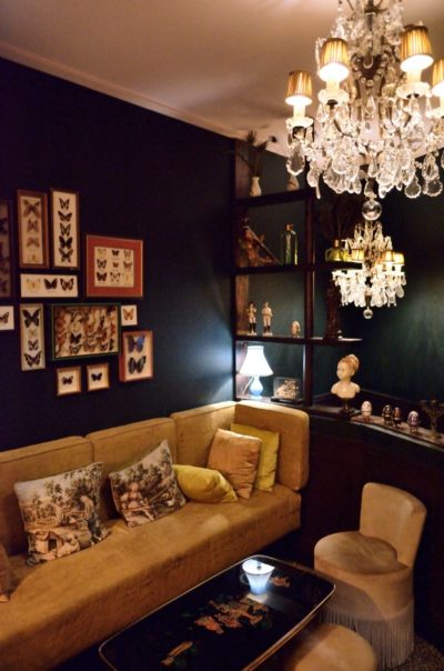 Le salon cosy de l'hôtel La Mondaine avec un canapé en velours orné de coussins décoratifs. Des œuvres d'art en forme de papillons accrochées au mur et un lustre aux détails en cristal projettent une lumière chaleureuse. Une table d'appoint avec un buste décoratif et divers bibelots ajoute du charme à la décoration éclectique, rappelant le style photographique parisien de Thomas Duclert.