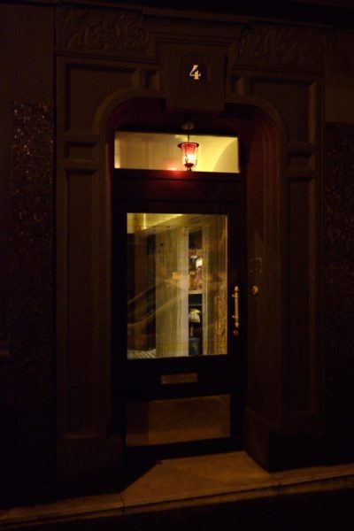 La porte d'entrée sombre et décorée de l'hôtel La Mondaine avec un panneau en verre, affichant le chiffre 4 au-dessus. La porte est éclairée par une lumière rouge chaude provenant d'une petite lanterne suspendue au-dessus. Le photographe Thomas Duclert a capturé cette scène parisienne enchanteresse où l'intérieur, faiblement visible à travers la vitre, révèle un escalier.