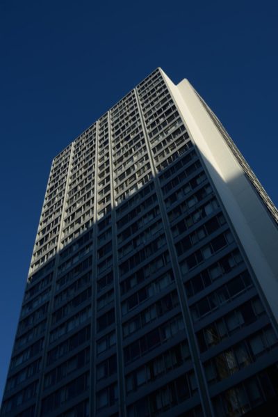 Un gratte-ciel moderne et haut, doté de nombreuses fenêtres, se détache sur un ciel bleu profond. Le bâtiment projette une ombre sur sa surface, créant un contraste entre les zones claires et sombres. Prise en contre-plongée, l'architecture évoque le style épuré souvent observé dans les créations parisiennes de Thomas Duclert.