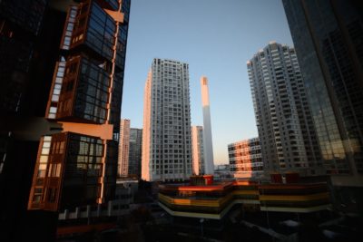Un paysage urbain avec des immeubles de grande hauteur au coucher du soleil, projetant une chaude lueur orange sur leurs façades. Les styles architecturaux modernes sont prédominants, avec le plus haut bâtiment au centre, entouré d'autres gratte-ciel et de structures aux fenêtres réfléchissantes, capturant un moment qui pourrait inspirer n'importe quel photographe à Paris.