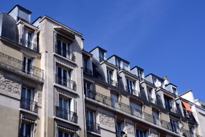 Vue en perspective d'immeubles d'appartements parisiens classiques sous un ciel bleu clair, capturée avec un souci du détail qui rappelle un photographe de mode comme Thomas Duclert. Les immeubles présentent des pierres ornementées, des balcons en fer forgé et de nombreuses lucarnes au dernier étage, certaines ornées de volets rouges ou blancs.