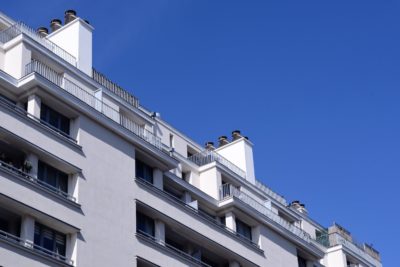 Une vue rapprochée d'un immeuble moderne à plusieurs étages avec des murs blancs et plusieurs balcons, sur fond de ciel bleu clair, capture l'essence de l'architecture parisienne. Le bâtiment dispose de plusieurs fenêtres et de cheminées sur le toit, ainsi que de balustrades métalliques sur les balcons.