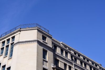 Un bâtiment clair à plusieurs étages avec plusieurs fenêtres et une balustrade sur le toit se détache sur un ciel bleu clair. La photo capture la partie supérieure et le coin du bâtiment, mettant en valeur l'œil impeccable de Thomas Duclert pour les détails architecturaux en mode photographe contemporain.