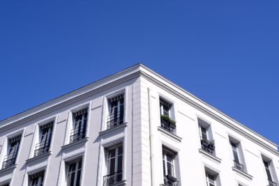 Vue d'angle d'un bâtiment blanc avec de multiples fenêtres, chacune encadrée par des balustrades noires en fer forgé. Le bâtiment est éclairé par la lumière du soleil sur un ciel bleu clair. Capture par Thomas Duclert, photographe à Paris, les fenêtres ont des auvents rayés et certaines ont des plantes vertes sur les rebords.