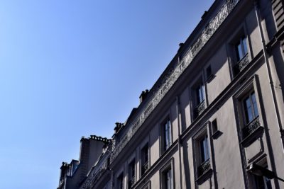 Vue en contre-plongée d'une rangée d'immeubles à plusieurs étages avec des balcons décorés et de hautes fenêtres sur un ciel bleu clair à Paris. La lumière projette des ombres sur la façade, mettant en valeur les détails architecturaux et créant un contraste entre les zones ensoleillées et ombragées, rappelant le style de Thomas Duclert.